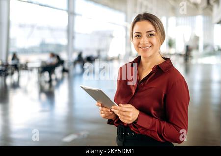 Portrait of positive beautiful intelligent caucasian business lady, recruiter, broker, company employee, in elegant clothes, holding a tablet in her hands, stand in the office, looks at camera, smiles Stock Photo