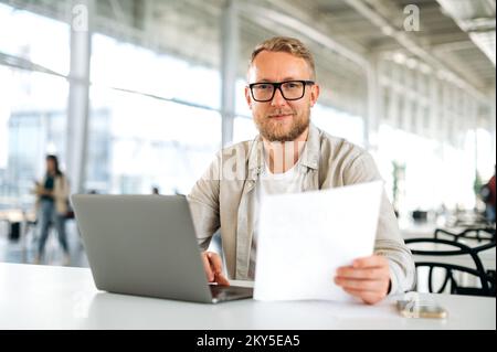 Friendly motivated successful caucasian man, with glasses, IT specialist, company seo, manager, freelancer, sits at a desk with a laptop in coworking, works with documents, looks at camera, smiles Stock Photo