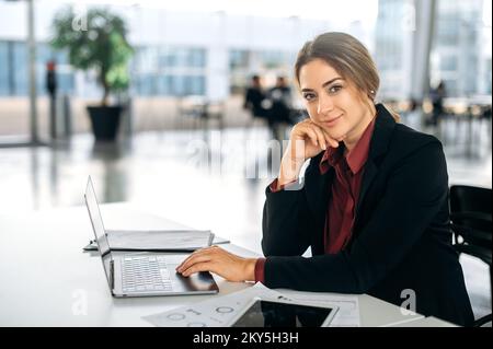 Photo of a gorgeous successful confident caucasian business woman, hr manager, broker, company employee, in elegant clothes, sitting at a workplace in the office, looks at camera, smiles friendly Stock Photo