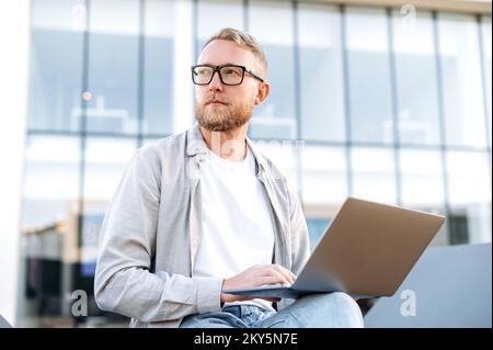 Serious focused caucasian bearded man with glasses, in casual clothes, sits outdoors near the office with a laptop, works on a project, looks away thoughtfully, plans a strategy, thinking over idea Stock Photo