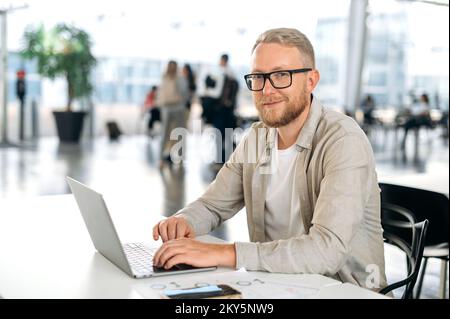Photo of a positive attractive clever caucasian business man, with glasses, executive, programmer, company seo, manager, sits at a desk with a laptop in coworking center, looks at camera, smiling Stock Photo