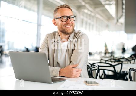 Positive smart successful caucasian man, with glasses, in stylish casual wear, IT specialist, company seo, freelancer, sits at a desk with a laptop in coworking, looks away, smiles happily Stock Photo