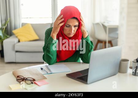 Concerned mature islamic woman using laptop, reading negative news online and touching head, sitting at home Stock Photo