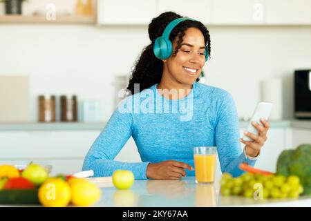 Happy black lady in sportswear using smartphone and wireless headphones, listening music in kitchen Stock Photo