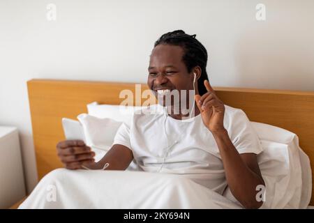 Cheerful Black Man Relaxing In Bed With Smartphone And Earphones Stock Photo