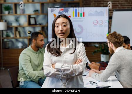 Confident happy smiling diverse asian woman, startup team leader, standing against background of mixed race office workers or business people with Stock Photo