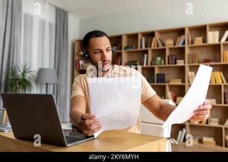 Focused arab man in wireless headphones having video call on laptop, working with documents at home in living room Stock Photo