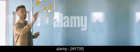 Millennial businessman putting sticky notes on glass wall in office, wide banner Stock Photo