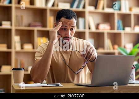 Sad tired arab male freelancer working on laptop and touching head, holding glasses, looking at computer screen Stock Photo