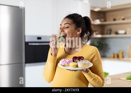 Smiling hungry young black pregnant female with big belly eats sweets, hold plate with cookies Stock Photo