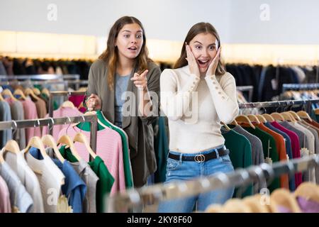 Amazed female holding warm clothes on hangers and showing her friend a discount during shopping in a store Stock Photo