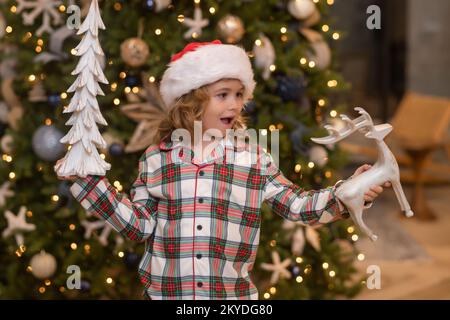 Christmas toys. Amazed kid holds a Christmas ball. The boy rejoices in the holiday, Christmas, New Year. Kid stands against the Christmas tree. Stock Photo