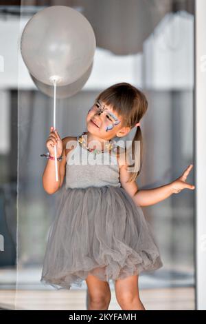 A girl in an elegant dress with a silver ball and face painting a butterfly poses beautifully like a doll. The child came to the birthday in the nursery High quality photo. Stock Photo