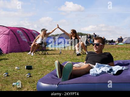 Yes to the fest. a young man relaxing on a blow up mattress outdoors while two friends high five in the background. Stock Photo