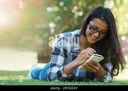 Jotting down her thoughts. A young woman lying on the grass writing in a notebook. Stock Photo
