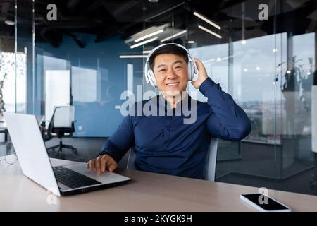 Portrait of happy asian worker in modern office, man looking at camera and smiling, boss programmer in shirt using headphones to watch video and listen to music at work. Stock Photo