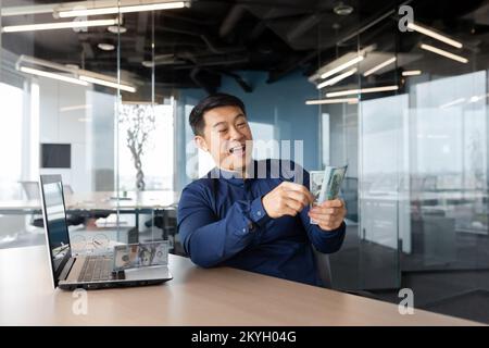 Happy and successful businessman counting money cash inside office, investor satisfied with achievements holding dollar bill in hands smiling working with laptop. Stock Photo