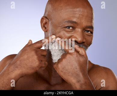 Face, beauty and skincare with a senior black man checking his skin for a pimple or zit in studio on a purple background. Portrait, wellness and Stock Photo