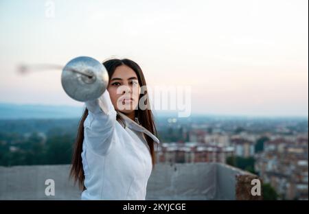Portrait of young woman fencer wearing mask, white fencing costume and practicing outdoors. Stock Photo