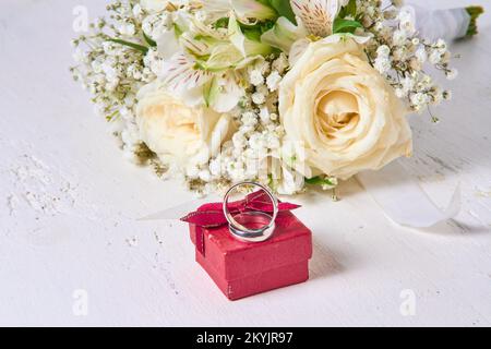Two white gold wedding rings, different sizes, lie on a red box and a bride's bouquet on a white wooden background. Stock Photo