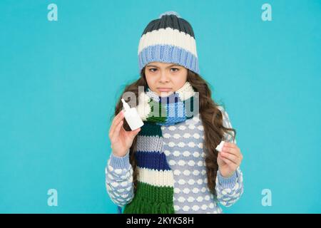 unhappy kid girl has runny nose and hold nasal spray in knitwear on blue background, coronavirus Stock Photo