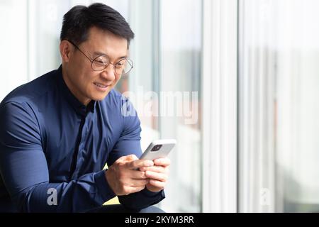 Closeup photo of a successful Asian businessman using the phone, a programmer in a shirt and glasses near a window inside an office messaging on social networks, using an application on a smartphone. Stock Photo