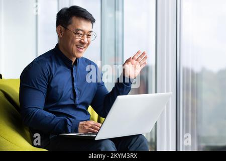 Video call, successful asian businessman near window in office talking remotely, online meeting with colleagues, programmer waving at laptop camera greeting gesture. Stock Photo