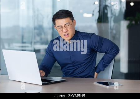 Sick overworked man at work, businessman has severe back pain, asian man holding hand on side massaging muscle, boss in shirt and glasses at work inside office using laptop. Stock Photo