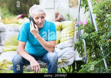 Elderly man talking on a cell phone while sitting on a garden swing Stock Photo