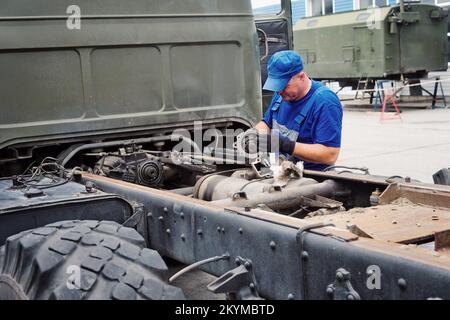 Mechanic in overalls and baseball cap repairs tractor or truck on summer day.. Front view. Professional assistance in maintenance of heavy equipment. Authentic workflow.. Stock Photo