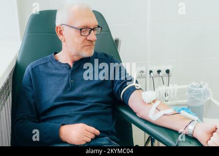 Donor donates blood. Elderly man of 50-60 years old in glasses sits in chair in medical clinic and donates blood from vein. Authentic scene. Volunteer donates blood to save life.. Stock Photo