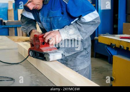 Professional woodworker grinds wooden bar on workbench in carpentry workshop with tool. Middle-aged worker in overalls processes wood with electric grinder. Real workflow.. Stock Photo