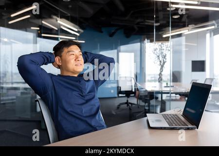 Asian man finished work well, businessman resting in office satisfied with achievement throw hands behind head dreaming, man working inside office using laptop at work. Stock Photo