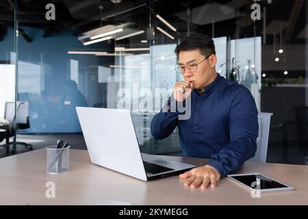 Asian man coughing at work in office, businessman working sick at workplace man using laptop, mature boss in glasses having strong cough. Stock Photo