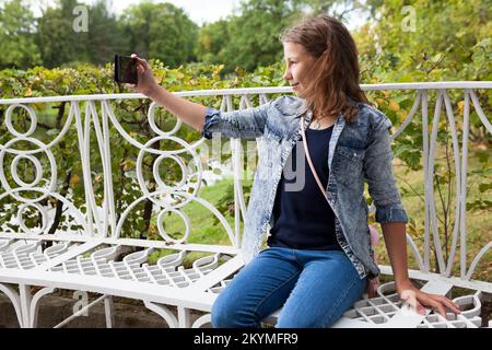 Teenage girl taking a selfie with her smartphone while sitting on a park bench Stock Photo