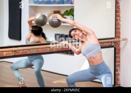 Slim young brunette with ball bending aside and doing plie squat near handrail and mirror during ballet rehearsal in studio Stock Photo