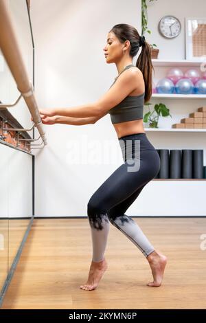 Full body side view of slim female ballet dancer in activewear performing releve exercise on barre during rehearsal in studio Stock Photo