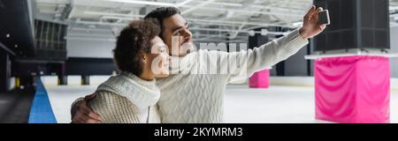 Positive interracial couple taking selfie on smartphone on ice rink, banner Stock Photo