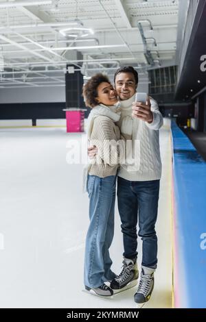 Young interracial couple in ice skates and warm clothes taking selfie on rink Stock Photo