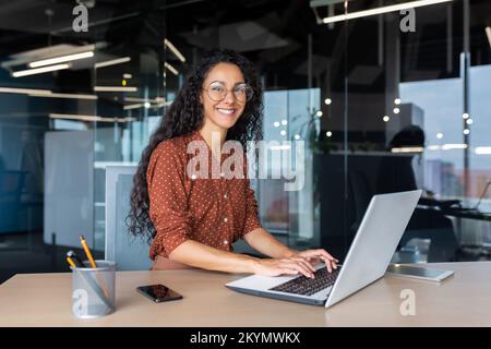 Young beautiful hispanic woman working inside modern office, businesswoman smiling and looking at camera at work using laptop. Stock Photo