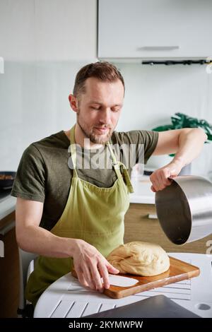 Making pastry at home kitchen concept. Bearded male chef baker in green apron getting out dough from metal bowl on a wooden cutting board using plastic spatula. High quality image Stock Photo