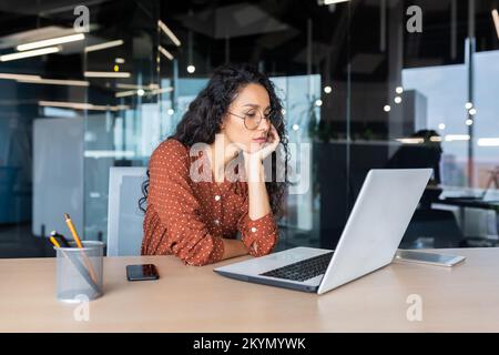Sad grumpy Latin American woman working inside the office, business woman is bored looking at the laptop screen. Stock Photo