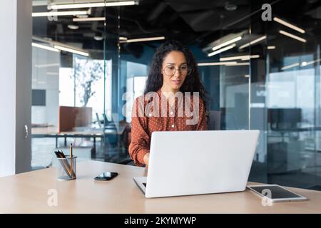 Cheerful and successful indian woman programmer at work inside modern office, tech support worker with laptop typing on keyboard smiling. Stock Photo