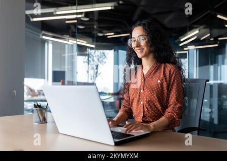 Cheerful and successful indian woman programmer at work inside modern office, tech support worker with laptop typing on keyboard smiling. Stock Photo