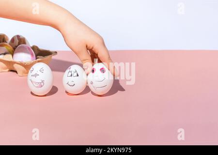 The child paints white eggs and puts them on a pink and white background. Funny and romantic faces drawn with a marker. Easter holiday concept. Stock Photo