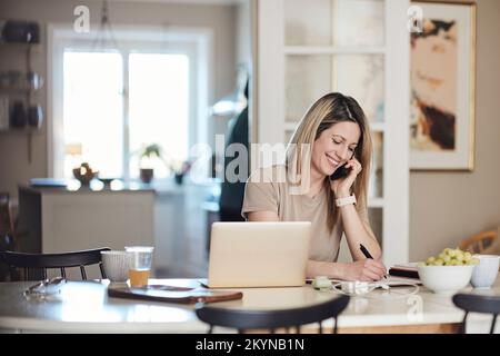 Happy freelancer taking down notes while talking on smart phone at home Stock Photo