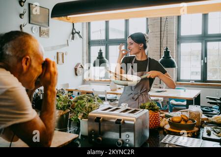 Male photographer taking picture of female chef gesturing while holding cooking pan at studio kitchen Stock Photo