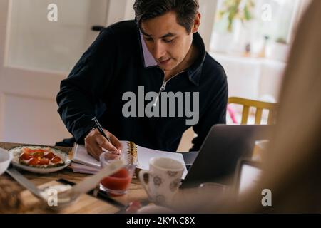Young male freelancer taking down notes while talking on smart phone at dining table Stock Photo