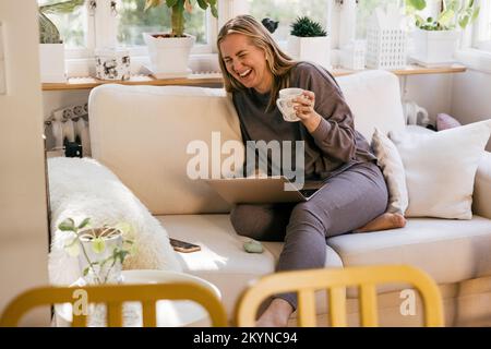 Happy female freelancer with laptop holding mug while sitting on sofa at home Stock Photo