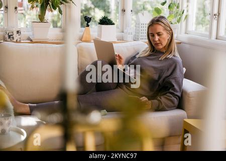 Blond female freelancer with wireless technologies reclining on sofa while working from home Stock Photo